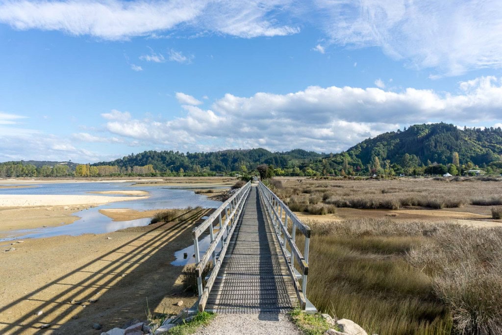 A passenger bridge in Marahau leading to the start of the Abel Tasman Coastal Track, with the Barn Cabins and Campground in the distance in the upper right corner of the frame, and a tidal ocean to the left.