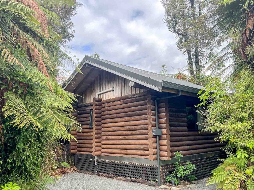 The exterior of a wooden cabin "treehouse" at Rainforest Retreat accommodation in Franz Josef, with ferns and greenery surrounding the building.