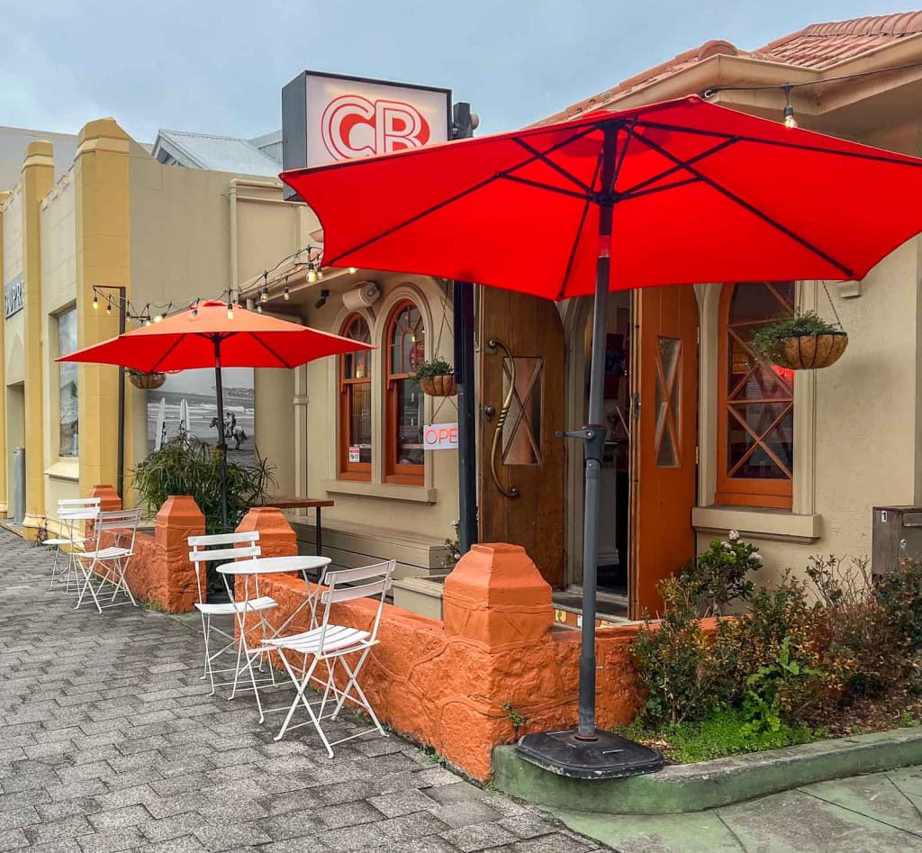 The exterior of Community Burgers in Napier, with outdoor tables and red umbrellas set up in the front courtyard.