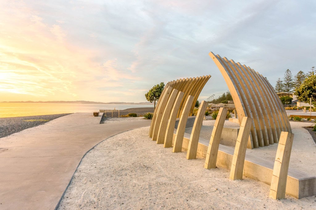 A sculpture resembling a whale bone along Marine Parade in Napier, with sunrise lighting over the ocean in the background.