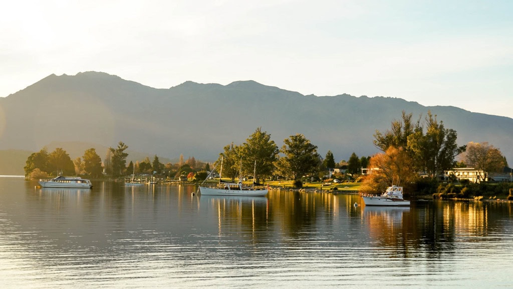 Lake Te Anau at sunset during golden hour lighting, with boats docked on the lake and mountains in the background.