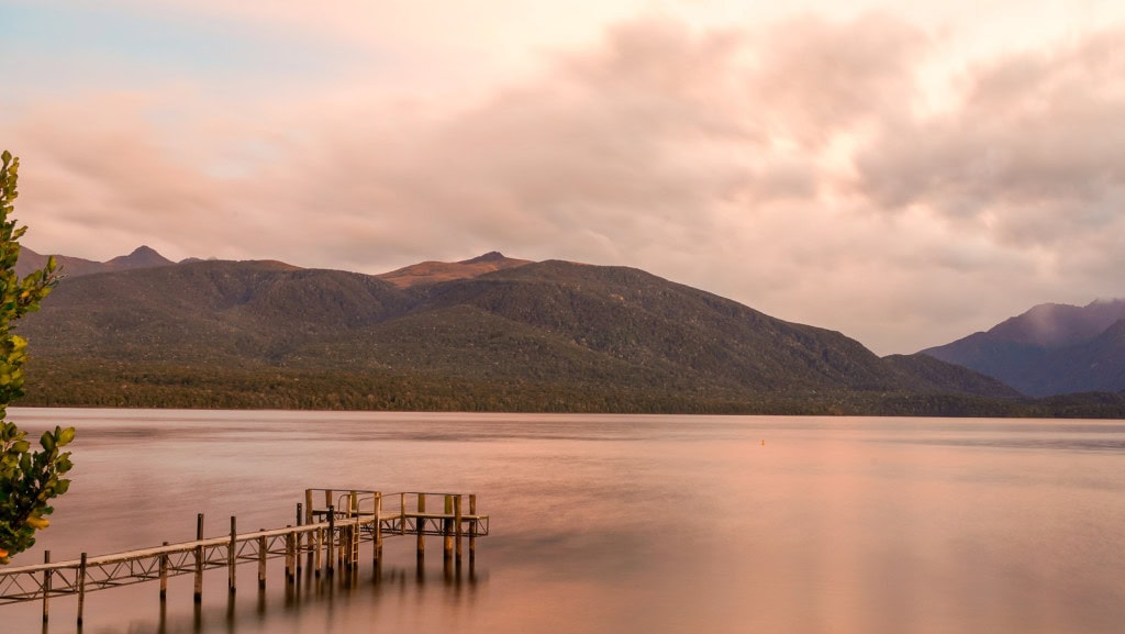The Marakura Wharf in Te Anau at sunrise, with bush-covered mountains in the background and a pinky tint to the lake and sky.
