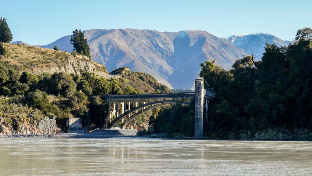 The Rakaia Gorge Bridge with the Rakaia River in the foreground and mountains in the backdrop.