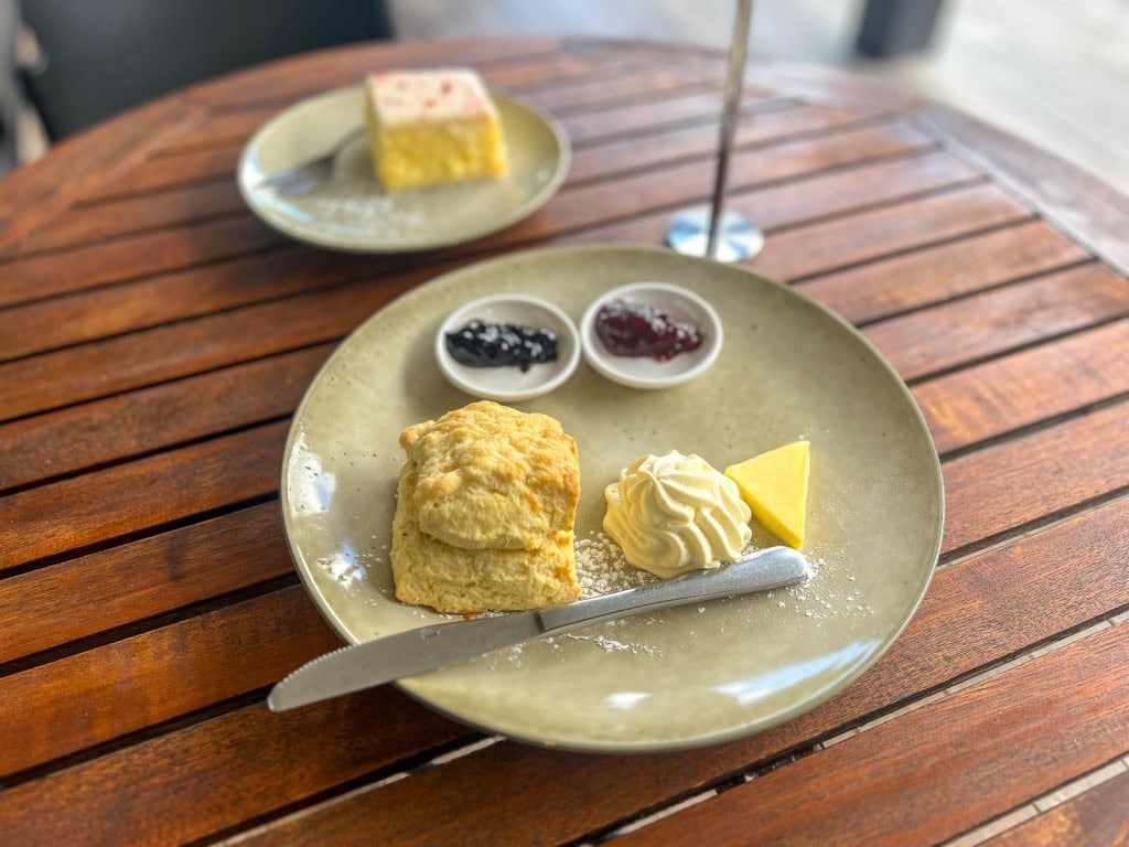 A scone with clotted cream, butter, and jam on plate on a wooden outdoor table at Barker's of Geraldine, with a custard slice on a plate behind it.